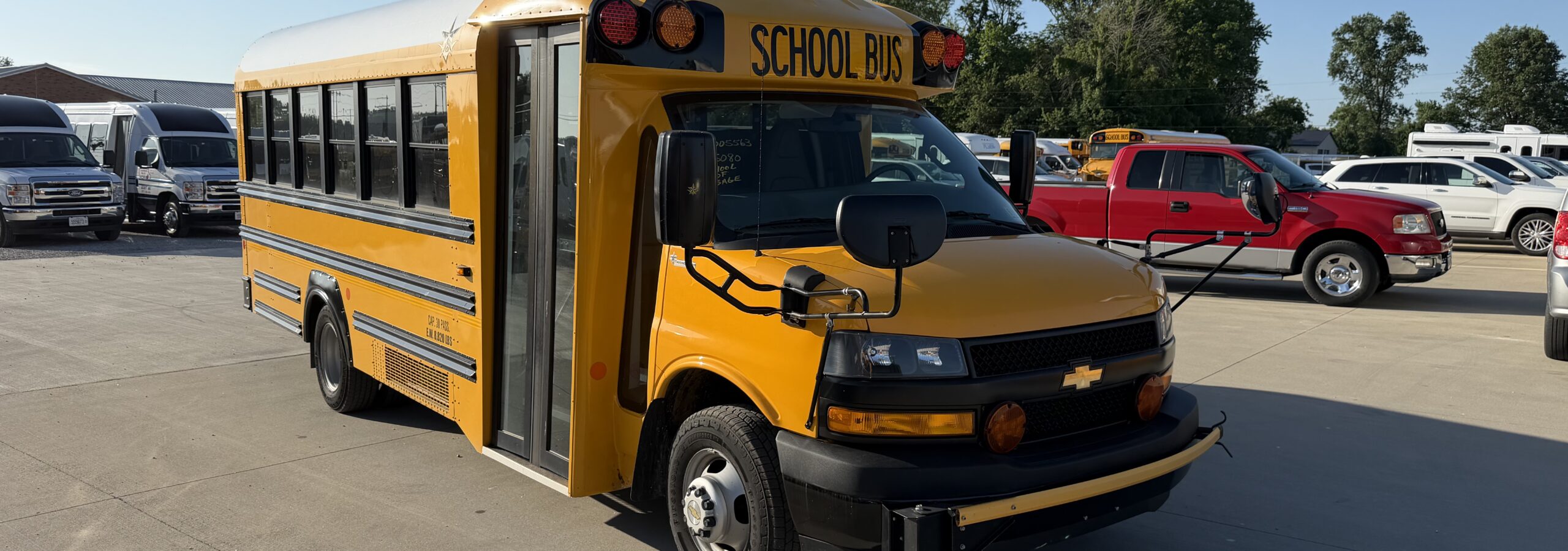 Yellow school bus parked in a lot with other vehicles and trees in the background, under a clear blue sky.