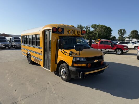 Yellow school bus parked in a lot with other vehicles and trees in the background, under a clear blue sky.