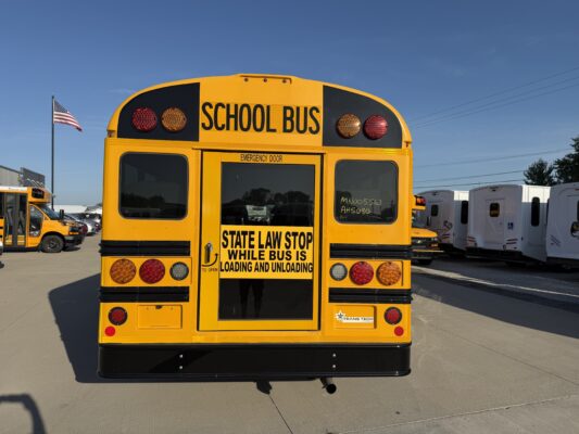 Back of a yellow school bus with emergency door, stop sign text, and red/amber lights; parking lot with more buses and an American flag nearby