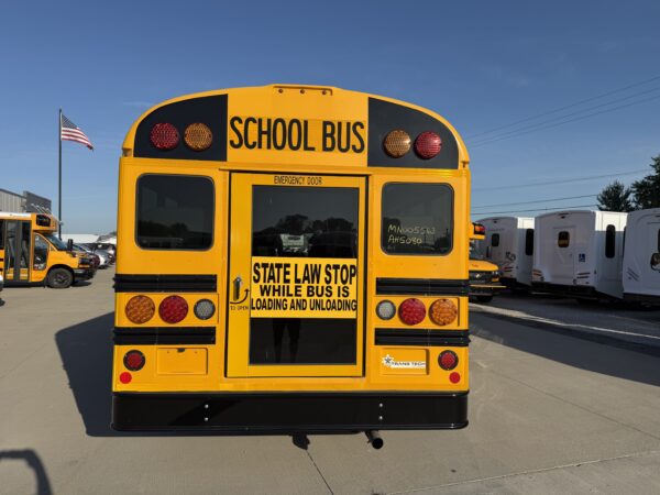 Back of a yellow school bus with emergency door, stop sign text, and red/amber lights; parking lot with more buses and an American flag nearby