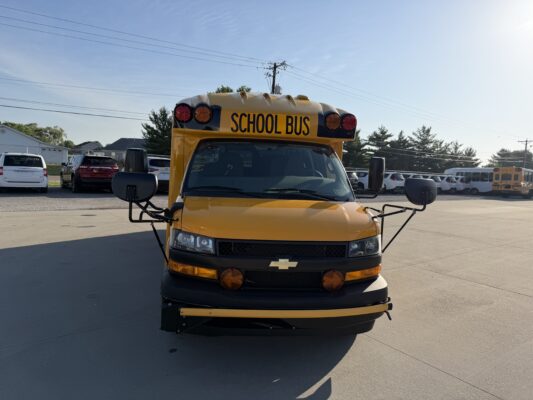 Front view of a yellow school bus with 'SCHOOL BUS' sign, parked in a lot with other vehicles and a clear blue sky above