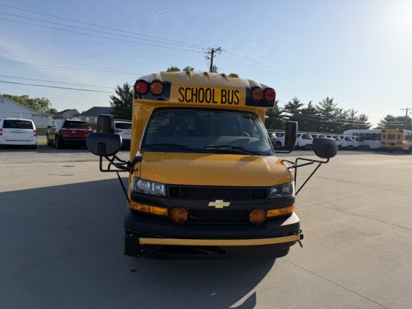 Front view of a yellow school bus with 'SCHOOL BUS' sign, parked in a lot with other vehicles and a clear blue sky above
