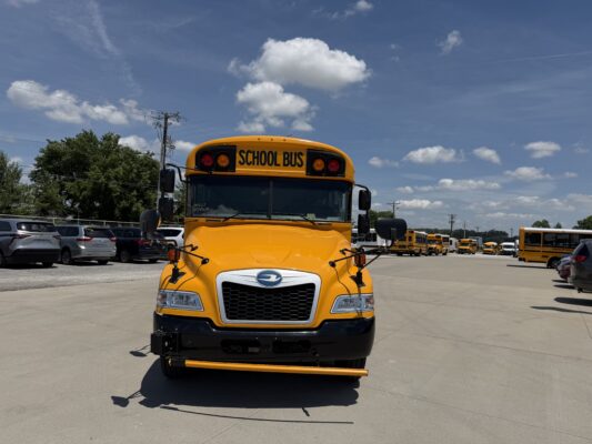 Front of a yellow school bus in a parking lot with other school buses in the background under a blue sky.