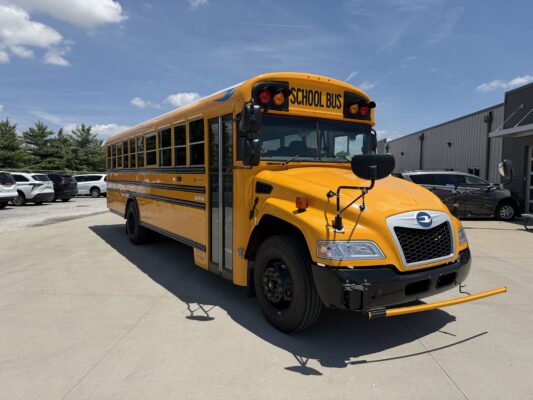 Front view of a yellow school bus parked in a lot, with a prominent 'SCHOOL BUS' sign on the top edge of the bus hood.