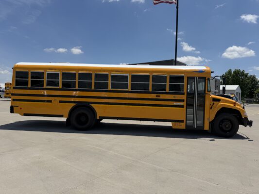 Side view of a bright yellow school bus parked in a lot under a clear blue sky, with a second bus and a flagpole in the background.
