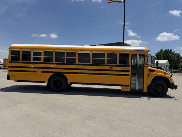 Side view of a bright yellow school bus parked in a lot under a clear blue sky, with a second bus and a flagpole in the background.
