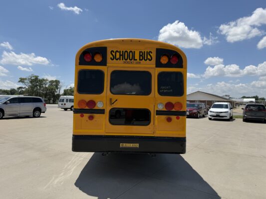 Back of a bright yellow school bus in a parking lot, with other vehicles and clear blue sky.