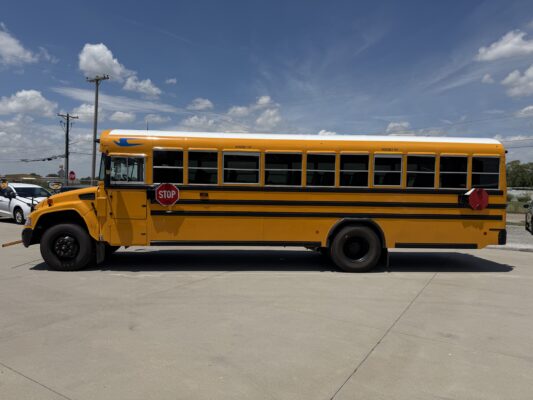 Yellow school bus parked in a lot with a stop sign and power lines under a blue sky with clouds.