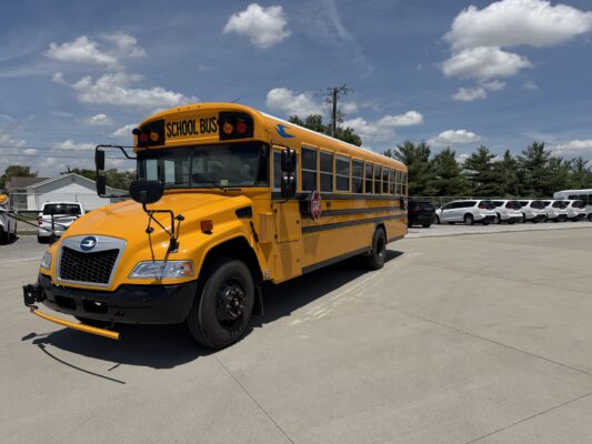 Close-up of a yellow school bus parked in a lot under a blue sky with white clouds, stop sign visible on the side edge.