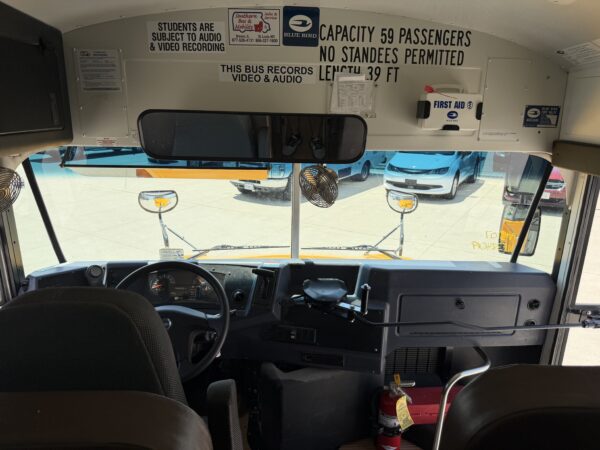 Front interior of a school bus: dashboard and steering wheel, overhead safety signs, and a mounted first aid kit on the wall.