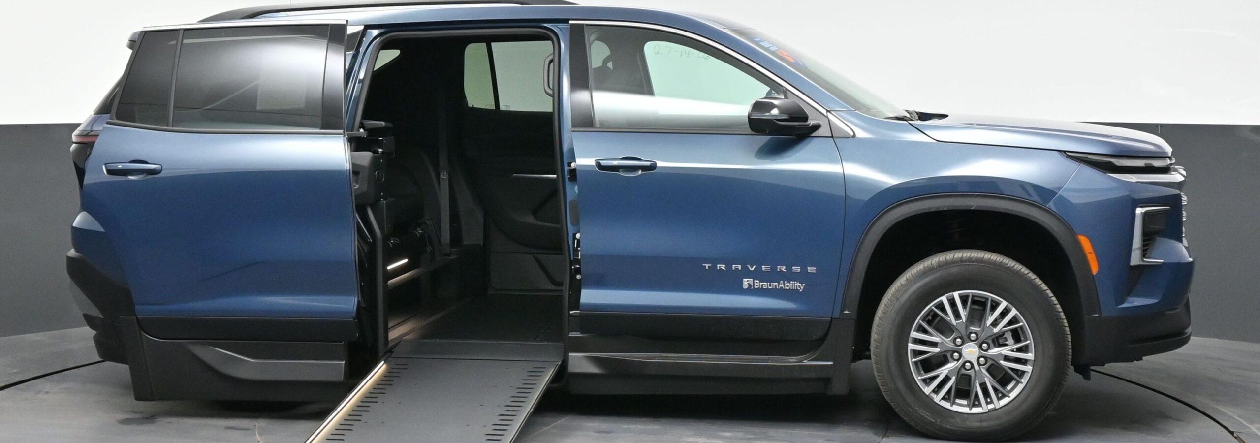 Blue SUV with a side-entry wheelchair ramp extended from the rear passenger area, in a showroom (BraunAbility branding visible).