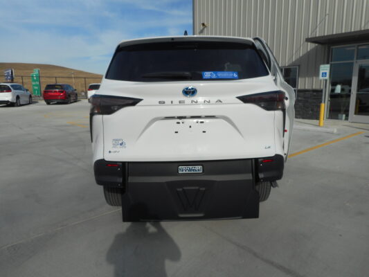 Rear view of a white Toyota Sienna minivan parked in a lot beside a modern building, with a handicap parking sign visible nearby.