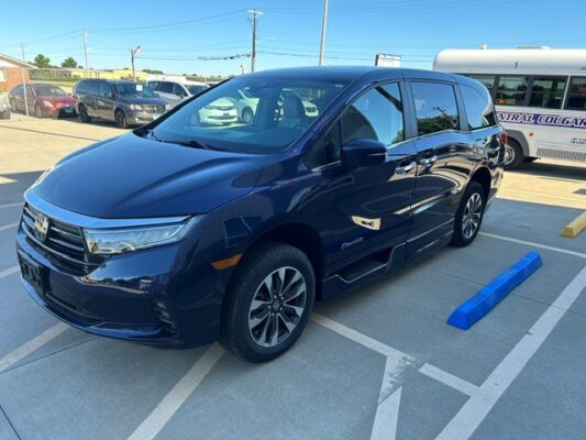 Front-left view of a dark blue minivan parked in a parking lot, with a blue curb block beside it and a school bus in the background.