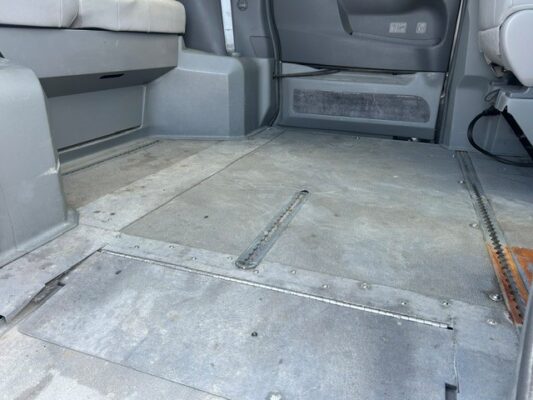 Interior of a vehicle showing a flat metal floor with embedded rails for a wheelchair ramp; doorway and seating visible at the back.
