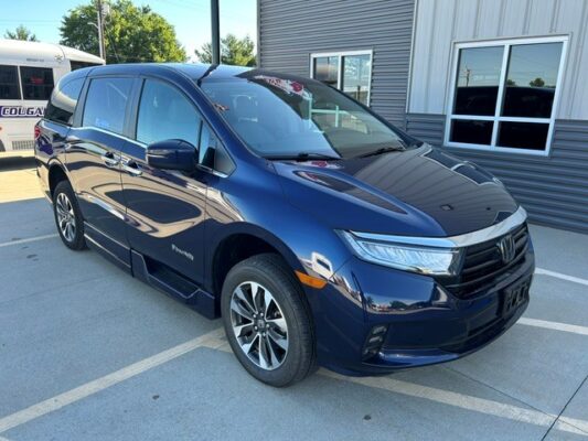 Front three-quarter view of a dark blue Honda minivan (Odyssey) parked in a dealership lot, hybrid badge visible on the side, with a building in the background.