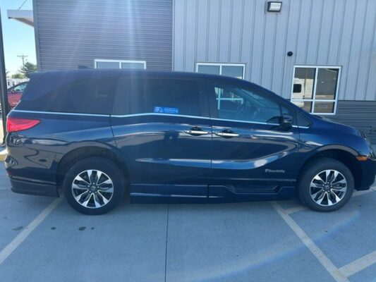 Blue minivan parked in a parking lot beside a metal industrial building, shown in full side view with sliding doors and alloy wheels.