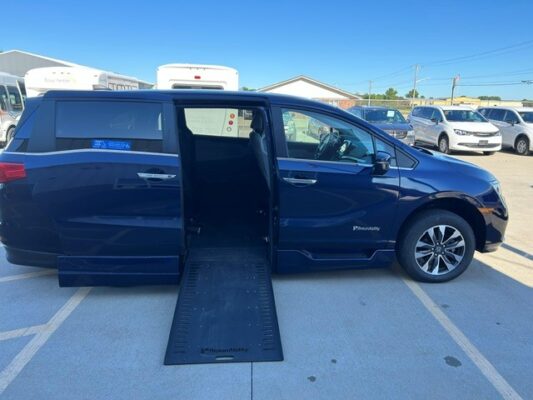 Blue minivan with an extended side ramp for wheelchair access in a parking lot. The rear and side doors are open, showing a lowered ramp and interior space ready for a wheelchair user.