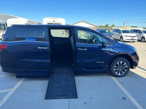 Ramp out (2) Blue minivan with an extended side ramp for wheelchair access in a parking lot. The rear and side doors are open, showing a lowered ramp and interior space ready for a wheelchair user.