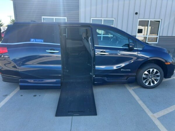 Ramp out Blue wheelchair-accessible van with its side ramp extended for entry, parked in a lot beside a metallic building.
