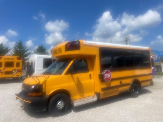 Front-right view of a yellow school bus with a stop sign extended on the side in a parking lot, other buses visible in the background.