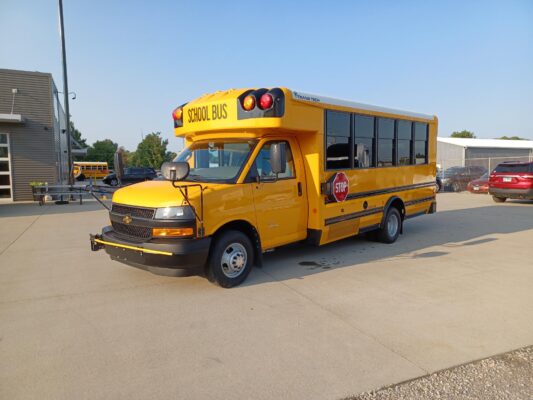 Front/side view of a yellow school bus parked in a lot, blue sky overhead, with another bus and buildings in the background, and a stop sign on the side.