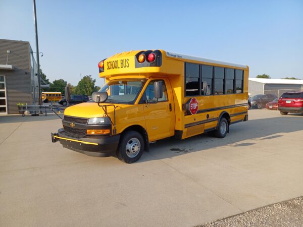 Front/side view of a yellow school bus parked in a lot, blue sky overhead, with another bus and buildings in the background, and a stop sign on the side.