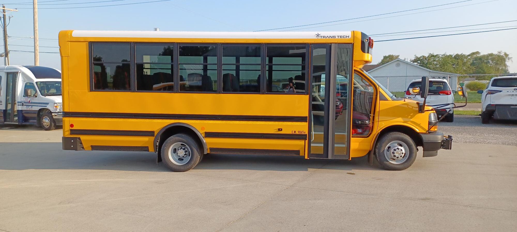 Yellow school bus parked in a lot, side view with open door and visible 'Trans Tech' logo near the front.