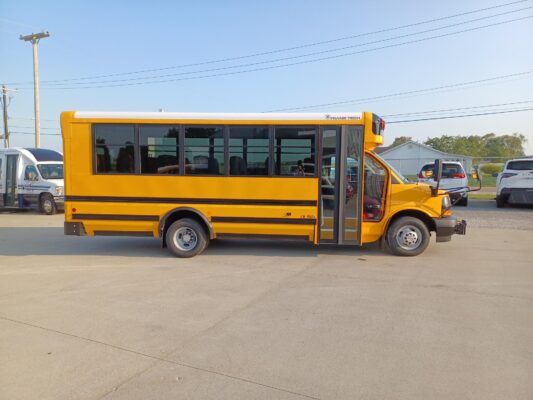 Yellow school bus parked in a lot, side view with open door and visible 'Trans Tech' logo near the front.