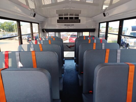 Interior of a school bus with gray seats and orange seat belts, aisle down the center and windows on both sides for daylight.