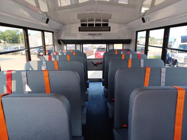 Interior of a school bus with gray seats and orange seat belts, aisle down the center and windows on both sides for daylight.