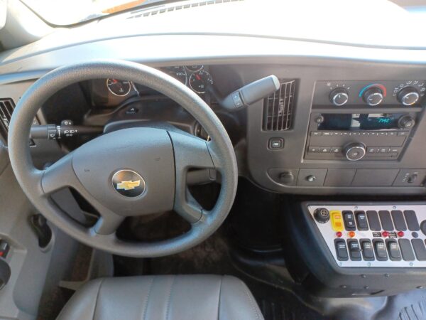 Interior of a Chevrolet car showing a gray steering wheel with Chevy emblem, instrument cluster, and a dashboard with climate controls and radio on the right side. A mounted control stalk extends from the steering column near the wheel. The windshield is bright outside.