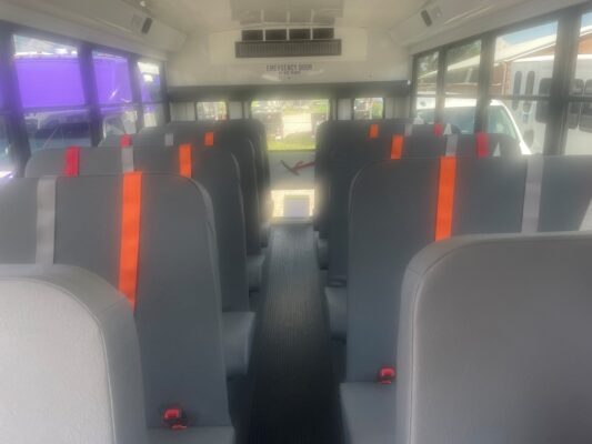 Interior of a bus with gray seats and orange seat belts, viewed from the back toward the front of the cabin edge-to-edge.