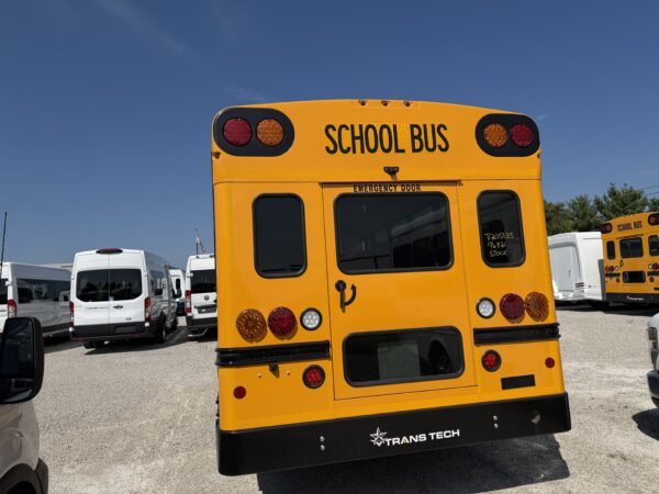 Rear of a bright yellow school bus with red and amber taillights, SCHOOL BUS label, in a parking lot with other buses nearby.