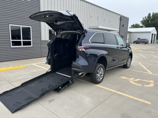 SUV with rear hatch open and a long black wheelchair ramp deployed to the side, parked in a lot by a gray industrial building—ready for loading or unloading a wheelchair user.