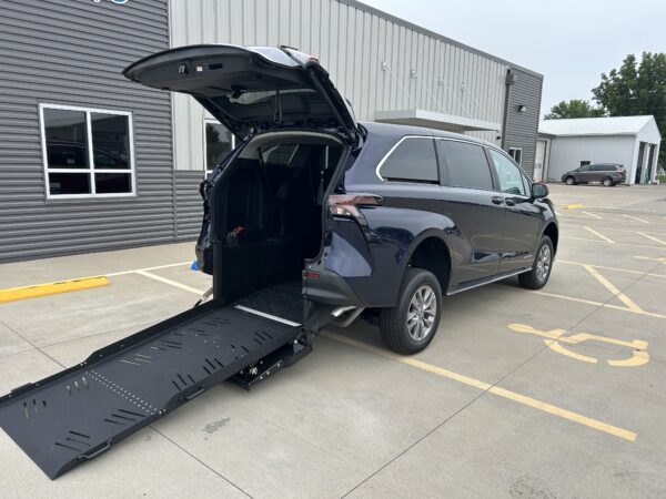 IMG_4974 SUV with rear hatch open and a long black wheelchair ramp deployed to the side, parked in a lot by a gray industrial building—ready for loading or unloading a wheelchair user.