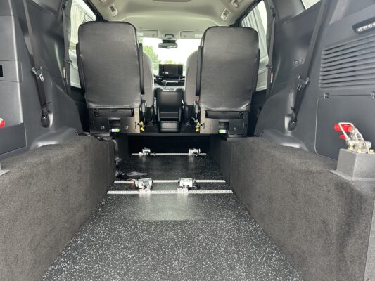 Rear interior of a passenger van with two rows of black seats and metal rail tracks along a speckled black floor viewed from the back arm—likely a modular seating setup.