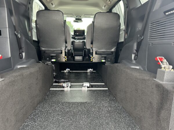 IMG_4975 Rear interior of a passenger van with two rows of black seats and metal rail tracks along a speckled black floor viewed from the back arm—likely a modular seating setup.