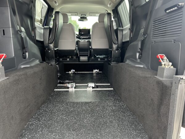 IMG_4976 Interior of a vehicle viewed from the back, showing front seats and an open cargo area with metal floor rails and speckled flooring.