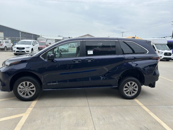 IMG_4977 Dark blue minivan in a parking lot with other cars and a chain-link fence in the background, showing a mobility vehicle with a side ramp area and 'Vantage Mobility' branding.