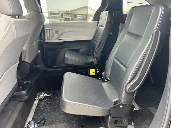 IMG_4979 Interior of a vehicle showing black leather captain's chairs in the second row, with one seat folded forward and a plastic door panel visible; outside parking lot seen through window.