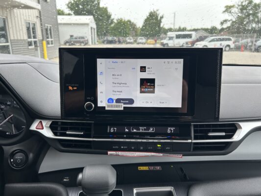 Dashboard touchscreen displaying radio options; raindrops on the windshield and a parking lot outside.