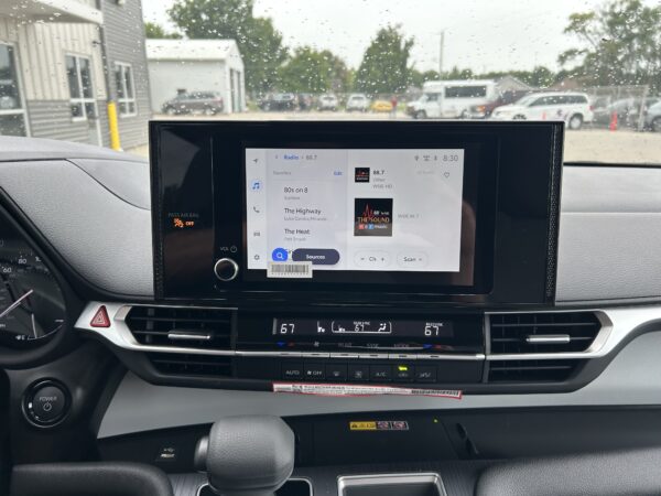 IMG_4980 Dashboard touchscreen displaying radio options; raindrops on the windshield and a parking lot outside.