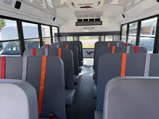 Interior of a empty school bus viewed from the back, gray seats with red, orange, and gray seatbelts along a central aisle near large windows