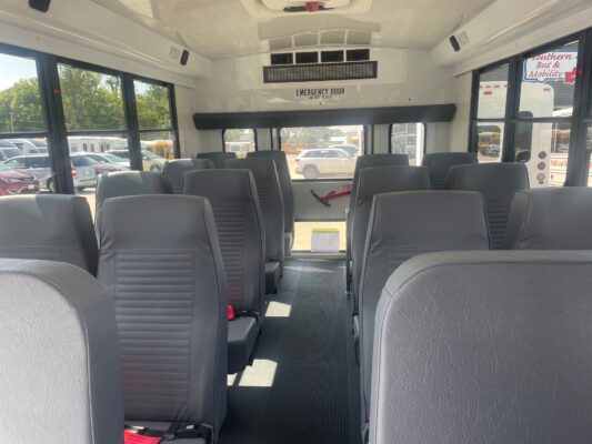 Interior of a parked bus with gray seats, a central aisle, and large windows showing cars outside; emergency exit area at the front/center.