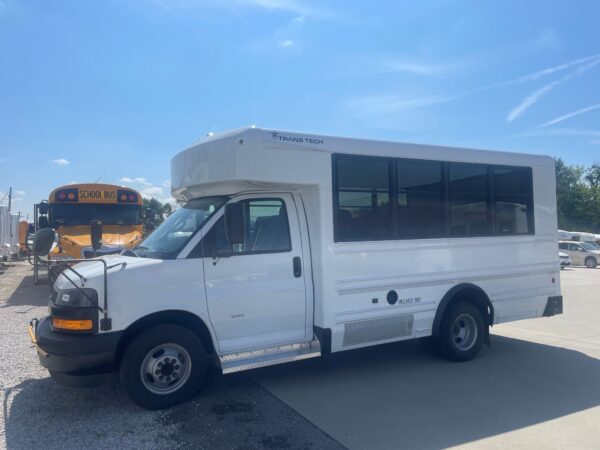 White shuttle bus with large windows and a Trans Tech logo on the side, parked in a lot with a yellow school bus in the background under a blue sky.