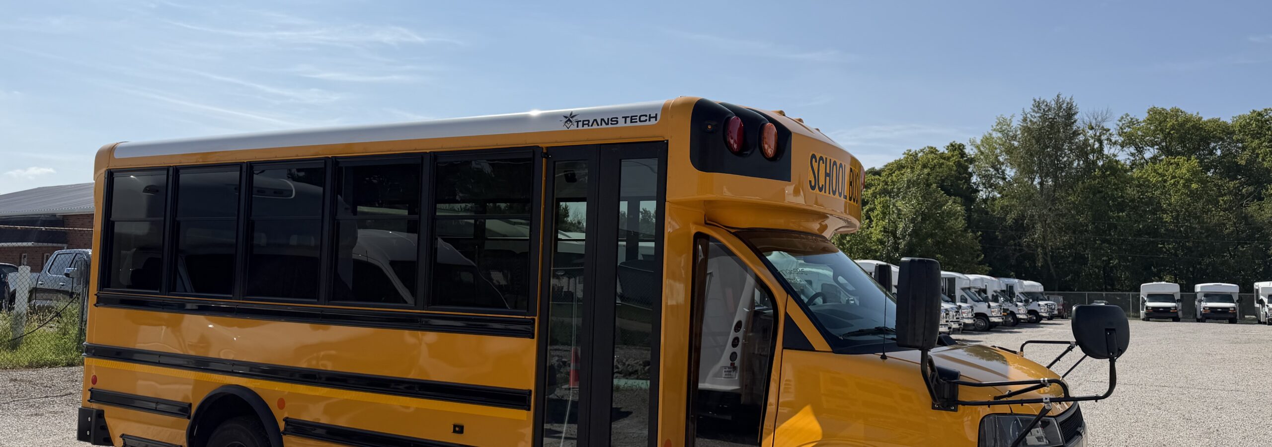Yellow school bus parked in a lot, showing the side and front with trees and blue sky in the background