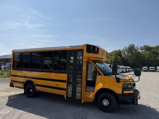 Yellow school bus parked in a lot, showing the side and front with trees and blue sky in the background