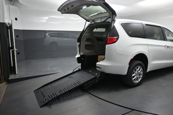 White minivan with the rear hatch open and a black wheelchair ramp extended to the floor, inside a light gray testing chamber, showcasing accessibility features.