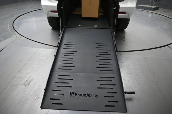 Wheelchair ramp extended from the back of a white car (BraunAbility) onto a gray metal surface, ready for loading a mobility device.