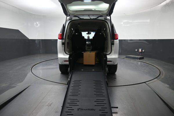 Rear view of a white wheelchair-accessible van with the ramp extended to the ground inside a white/gray studio, a cardboard box on the ramp near the rear seats.